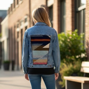 Puede incluir: Chaqueta vaquera azul claro con un diseño tejido colorido en la espalda. La chaqueta tiene un cuello clásico, mangas largas y cierres de botón. El panel trasero muestra un patrón geométrico en tonos de azul, marrón, naranja y crema.