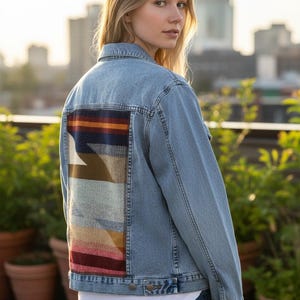 May include: Light blue denim jacket with a colorful geometric design on the back. The design features stripes of red, orange, blue, and brown. The jacket has a classic collar, button closures, and pockets.