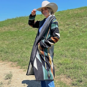May include: A woman wearing a white cowboy hat and a long, black, green, and brown patterned coat. She is standing on a dirt path with green grass on either side.