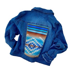 Könnte beinhalten: Blaue Jeansjacke mit farbenfrohem Design im Südweststil auf dem Rücken. Das Design zeigt geometrische Muster in Blau-, Braun- und Weißtönen. Die Jacke hat einen klassischen Kragen, Knopfverschlüsse und Taschen.
