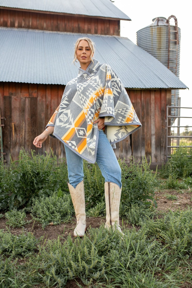 May include: A woman wearing a grey, yellow and white patterned poncho with a geometric design. She is wearing blue jeans and white cowboy boots. The woman is standing in a field of green grass.