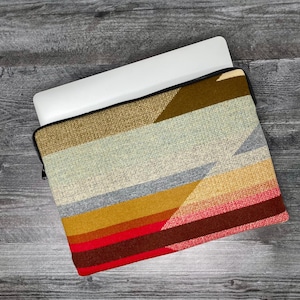May include: A laptop sleeve with a colorful geometric pattern. The sleeve is made of a woven fabric with stripes of red, orange, yellow, blue, and gray. The sleeve has a zipper closure.