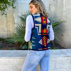 May include: A woman wearing a blue, red, yellow and white patterned waistcoat with a white long-sleeved shirt and light blue jeans.