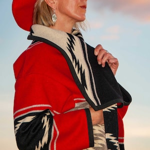 Puede incluir: Un sombrero rojo de ala ancha combinado con un abrigo largo que presenta un patr&oacute;n geom&eacute;trico audaz en rojo, negro y beige. El abrigo tiene un cuello chal ancho y ribetes negros en los bordes, mostrando un dise&ntilde;o de zigzag repetitivo.