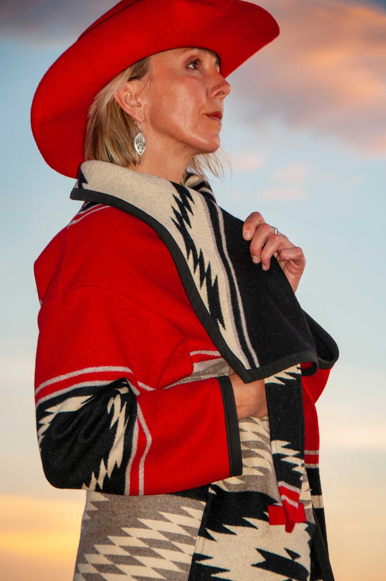 Puede incluir: Un sombrero rojo de ala ancha combinado con un abrigo largo que presenta un patr&oacute;n geom&eacute;trico audaz en rojo, negro y beige. El abrigo tiene un cuello chal ancho y ribetes negros en los bordes, mostrando un dise&ntilde;o de zigzag repetitivo.