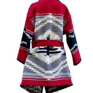 Puede incluir: Un abrigo de manga larga con un llamativo patr&oacute;n geom&eacute;trico en rojo, azul marino, gris y crema. La prenda incluye un cintur&oacute;n de tela roja a juego en la cintura, un cuello ancho y un dobladillo estructurado, exhibido sobre un maniqu&iacute;.