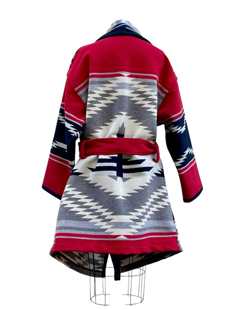 Puede incluir: Un abrigo de manga larga con un llamativo patr&oacute;n geom&eacute;trico en rojo, azul marino, gris y crema. La prenda incluye un cintur&oacute;n de tela roja a juego en la cintura, un cuello ancho y un dobladillo estructurado, exhibido sobre un maniqu&iacute;.