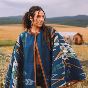 Può includere: Una donna indossa un poncho blu e marrone con un motivo geometrico. Il poncho ha un bordo marrone e una chiusura a bottone. La donna è in piedi in un campo con un edificio in legno sullo sfondo.
