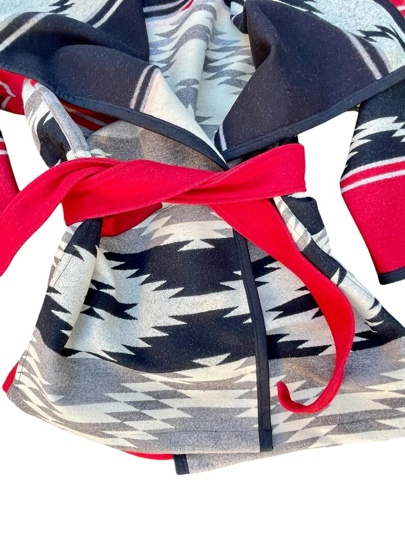 Puede incluir: Una chaqueta tipo envolvente con un patr&oacute;n geom&eacute;trico en negro, blanco y gris. La prenda incluye un cintur&oacute;n de tela roja s&oacute;lida atado a la cintura, mangas largas y un cuello ancho con ribete negro a lo largo de los bordes de los paneles de tela.