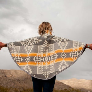 May include: A person wearing a grey and orange patterned poncho with a geometric design. The poncho is made of a soft, woven fabric and has a fringed edge. The person is standing in front of a mountain range.