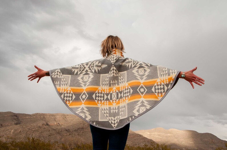 May include: A person wearing a grey and orange patterned poncho with a geometric design. The poncho is made of a soft, woven fabric and has a fringed edge. The person is standing in front of a mountain range.