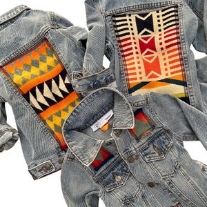 Puede incluir: Tres chaquetas vaqueras de lavado claro con paneles de tela tejida colorida en la espalda y uno en la parte delantera. Los paneles presentan patrones geométricos en tonos naranja, amarillo, rojo, negro y blanco. Las chaquetas tienen cierre de botón y bolsillos.