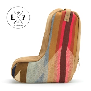 May include: A brown suede and patterned fabric footrest with a zipper on the side. The footrest is shaped like a boot and has a geometric pattern in shades of brown, pink, blue, and white. The footrest has a leather strap with a metal buckle on the top. The footrest is labeled "L7 AZ DESIGNS" in a circle with a star and arrows.
