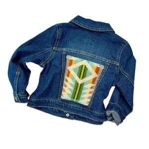 Puede incluir: Una chaqueta vaquera azul con un patrón geométrico colorido en la espalda. El patrón está en tonos de azul, verde, amarillo y marrón.