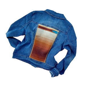 Könnte beinhalten: Eine blaue Jeansjacke mit einem bunten gewebten Patch auf der Rückseite. Der Patch zeigt einen Farbverlauf in Braun, Orange und Weiß.