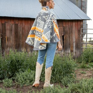 May include: A woman wearing a grey and orange patterned poncho with a geometric design. She is standing in a field with a barn and silo in the background. She is wearing blue jeans and white cowboy boots.