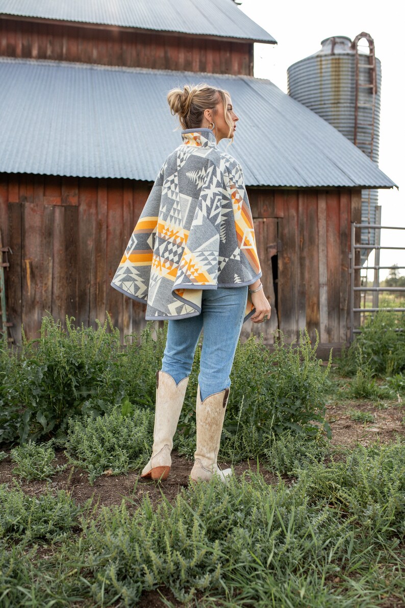 May include: A woman wearing a grey and orange patterned poncho with a geometric design. She is standing in a field with a barn and silo in the background. She is wearing blue jeans and white cowboy boots.