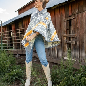 May include: A woman wearing a grey and yellow patterned poncho with a geometric design, blue jeans, and white cowboy boots. She is standing in front of a red barn.