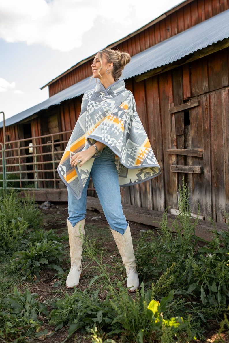 May include: A woman wearing a grey and yellow patterned poncho with a geometric design, blue jeans, and white cowboy boots. She is standing in front of a red barn.
