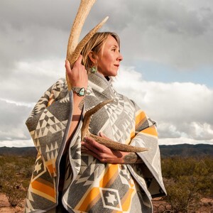 May include: A woman wearing a grey and yellow patterned poncho holds a large antler in front of her. She is standing in a desert landscape with a cloudy sky in the background.
