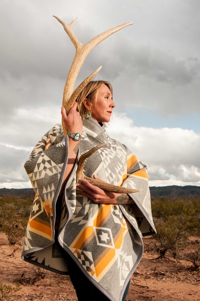 May include: A woman wearing a grey and yellow patterned poncho holds a large antler in front of her. She is standing in a desert landscape with a cloudy sky in the background.