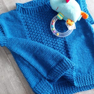 May include: A blue knitted jumper with a textured pattern. The jumper has long sleeves and a round neckline.