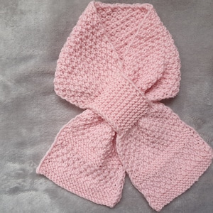 May include: A pink knitted scarf with a simple stitch pattern. The scarf is tied in a bow.