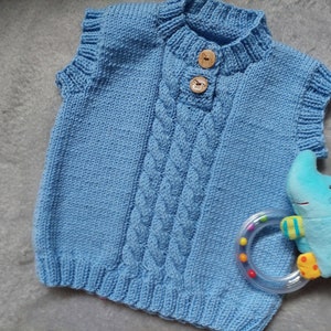 May include: A light blue knitted vest with a cable knit pattern and two wooden buttons. The vest has a high neck and sleeveless design.