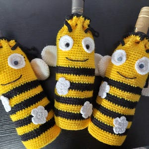 Puede incluir: Tres cubiertas de botella de vino de ganchillo con forma de abejorro. Cada cubierta es de rayas amarillas y negras con detalles blancos y tiene una cara de ganchillo con ojos saltones.