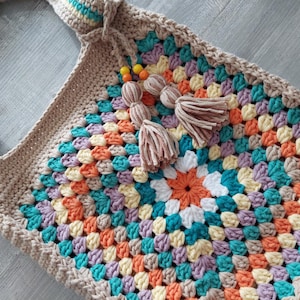 Könnte beinhalten: Handgefertigte Häkeltasche mit farbenfrohem Granny-Square-Design. Die Tasche hat eine beige Basis mit einem lebendigen Muster aus orangefarbenen, türkisfarbenen, gelben und lila Quadraten. Sie hat einen passenden beigen Riemen und ist mit Quasten verziert.