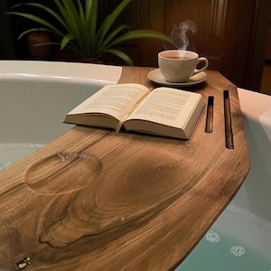 Puede incluir: Una bandeja de bañera de madera con un libro, una taza de té y un platillo. La bandeja tiene una hendidura tallada para una pastilla de jabón y ranuras para una tableta o teléfono. El libro está abierto y sale vapor del té.