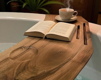 Peut inclure: Un plateau de baignoire en bois naturel avec un grain de bois apparent, une empreinte circulaire et deux fentes rectangulaires. Un livre ouvert est posé sur la surface à côté d'une tasse et d'une soucoupe en céramique blanche fumante.
