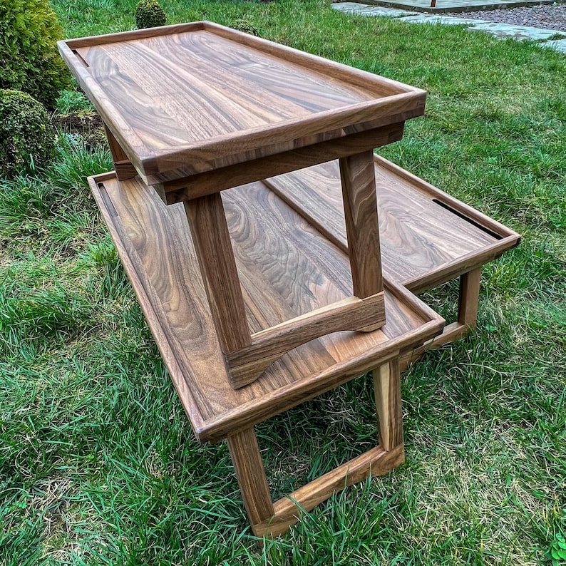May include: Two wooden serving trays with a natural wood grain finish are stacked on green grass. The trays have a rectangular shape with raised edges and are supported by angled legs. The wood has a warm, brown tone.