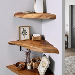 Może przedstawiać: Trzy drewniane półki narożne z naturalną krawędzią zamontowane na jasnoszarej ścianie. Półki zawierają elementy dekoracyjne, takie jak oprawiony nadruk paproci, małe lustro i drewniana miska. Widoczny jest również szalik i świeca.