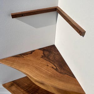 May include: A corner shelf made of natural wood, featuring a triangular design. The shelf has a live edge and is supported by two wooden brackets. The wood grain is visible, showcasing a mix of light and dark brown tones. The shelf is designed to fit into a corner.