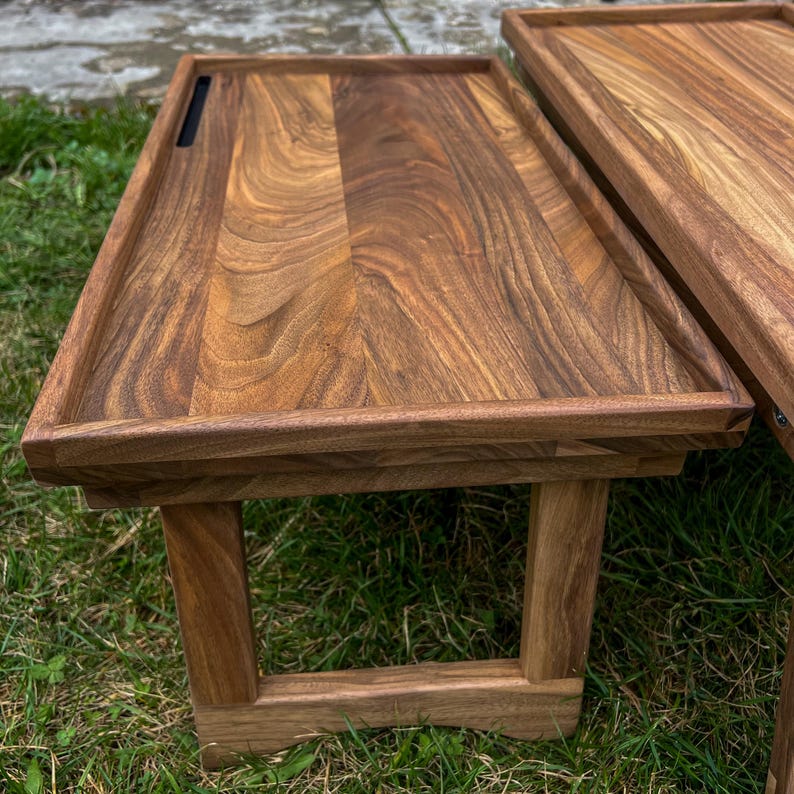 May include: Two wooden serving trays with a dark brown finish. The trays have a rectangular shape with raised edges and are supported by wooden legs. One tray has a small, rectangular cutout.