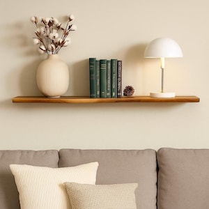 May include: A wooden shelf displays a white vase with cotton stems, several green books, a white lamp, and a pine cone. The shelf is mounted on a beige wall above a gray sofa with pillows.