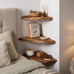 Peut inclure: Trois étagères d'angle en bois avec divers objets. L'étagère supérieure contient un panier tressé avec des lunettes et une petite bouteille marron avec des roseaux. L'étagère du milieu a des livres et une assiette décorative. L'étagère inférieure a un haut-parleur blanc, une tasse et un carnet.