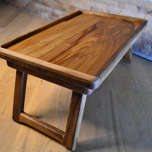 May include: A handcrafted wooden serving tray with a rectangular design. The tray features a raised border and angled legs, showcasing the natural wood grain. The warm brown tones of the wood create a rustic aesthetic, suitable for home decor.