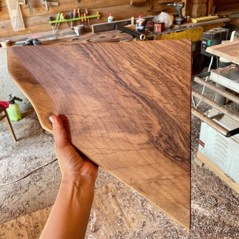 May include: A handcrafted, triangular wooden serving board with a natural wood grain pattern. The board has a curved edge on one side and is held by a hand. The wood has a warm brown color with darker streaks.