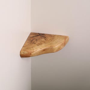 May include: A triangular corner shelf made from a single piece of light-colored natural wood. The shelf features a prominent live edge along the front curve and displays visible wood grain patterns, knots, and dark organic markings on its surface.