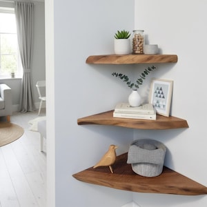 May include: Three wooden corner shelves on a white wall, each with decorative items. The top shelf has a potted plant and a jar of nuts. The middle shelf displays books and a framed picture. The bottom shelf features a wooden bird and a grey basket.