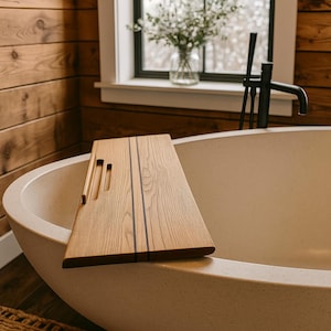 Puede incluir: Una bandeja de bañera de madera con acabado marrón claro y líneas oscuras. La bandeja tiene recortes para una tableta o libro y un asa. La bandeja descansa sobre el borde de una bañera ovalada beige.