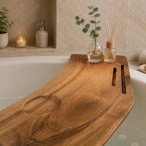 Puede incluir: Una bandeja de madera curva para bañera con acabado de veta natural, una hendidura circular para una taza o vela y dos ranuras rectangulares estrechas. La bandeja descansa sobre los bordes de una bañera blanca y sostiene un jarrón de vidrio.
