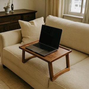 May include: A wooden laptop tray with a dark finish sits on a beige sofa. A laptop with a black screen rests on the tray. The tray has a rectangular top and angled legs, designed for use on a couch or bed.