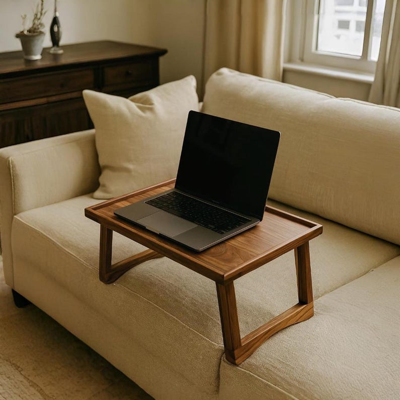 May include: A wooden laptop tray with a dark finish sits on a beige sofa. A laptop with a black screen rests on the tray. The tray has a rectangular top and angled legs, designed for use on a couch or bed.