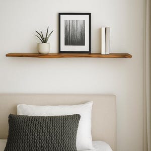 May include: A wooden floating shelf with a live edge, holding a framed black and white print, a potted plant, and two books. The shelf is mounted on a white wall above a bed with pillows.