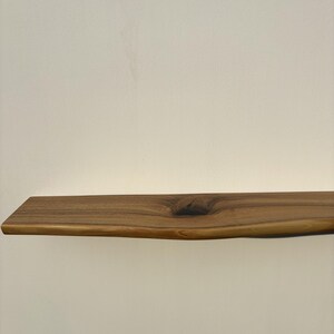 May include: A natural wood floating shelf with a live edge. The shelf is a warm brown color and features a knot in the wood. The shelf is approximately 30 inches long.