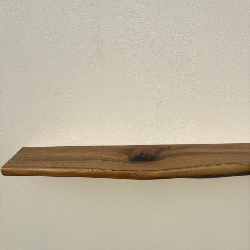 May include: A natural wood floating shelf with a live edge. The shelf is a warm brown color and features a knot in the wood. The shelf is approximately 30 inches long.