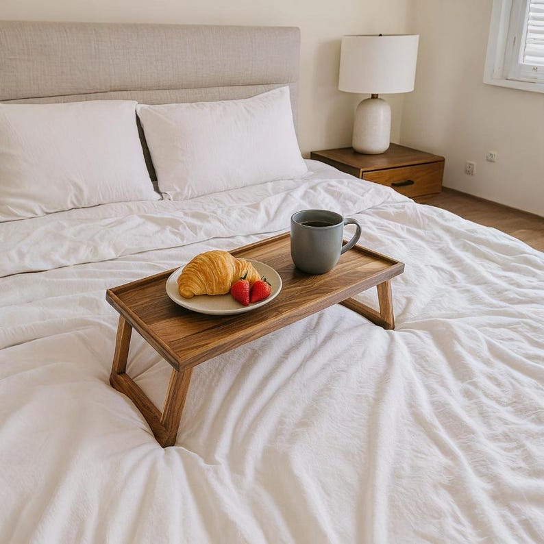 May include: A wooden bed tray with angled legs holds a plate with a croissant and two strawberries, and a gray mug. The tray sits on a bed with white linens, pillows, and a headboard. A lamp and nightstand are in the background.